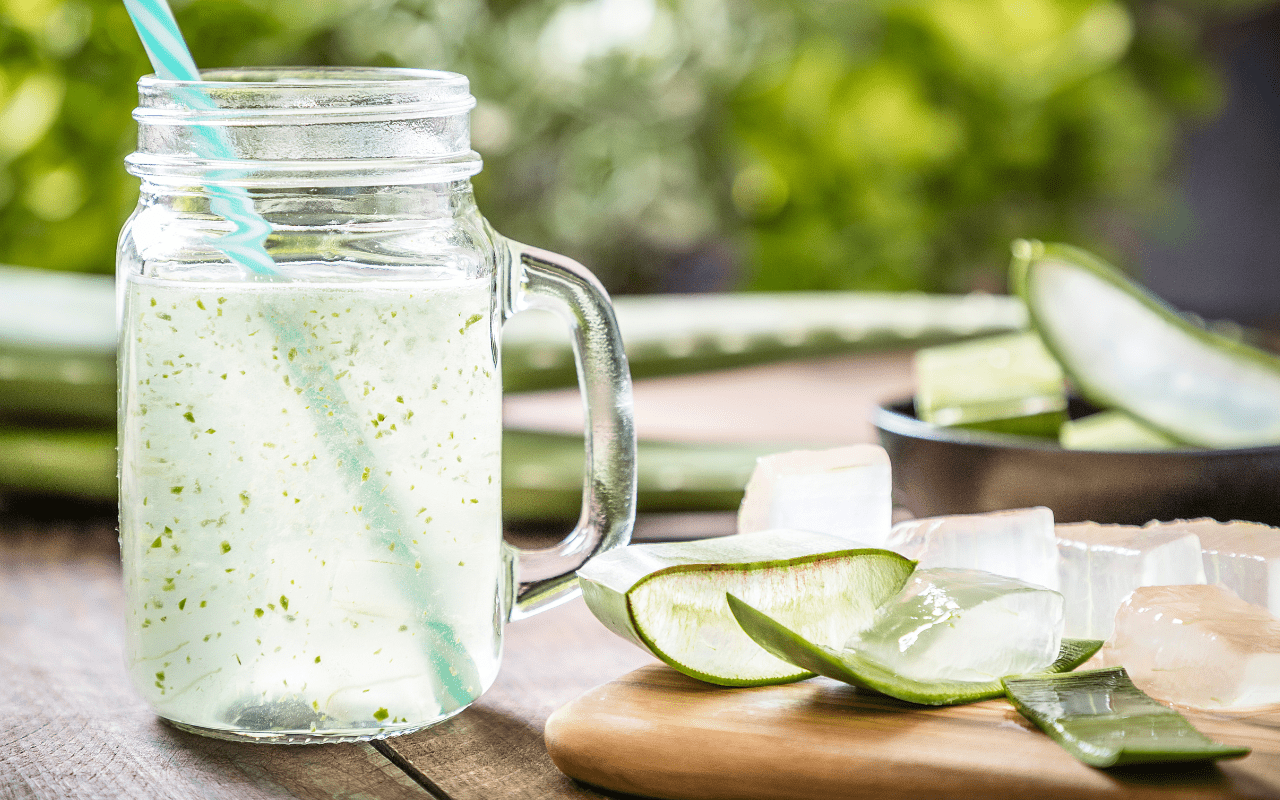 Sklenice aloe vera šťávy s ledem a čerstvě rozříznutými listy aloe na dřevěném prkénku