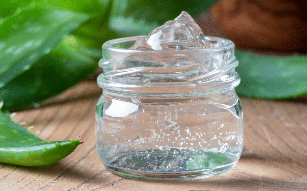 Skleněná dóza s čirým gelem z aloe vera na dřevěném stole s listy aloe v pozadí