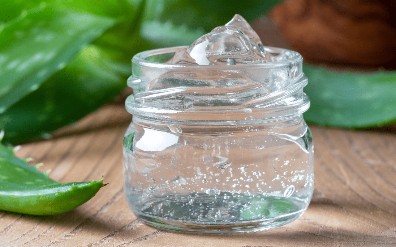 Skleněná dóza s čirým gelem z aloe vera na dřevěném stole s listy aloe v pozadí